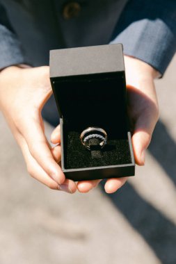 Eller, açık havada güneşli bir yerde parıltılı bir nişan yüzüğü içeren bir kara kutuyu gösteriyor..