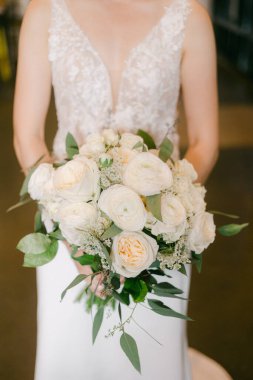 Bir gelin zarif bir şekilde durur, güzel bir buket beyaz gül ve yeşillik sergiler, neşe ve zarafet saçar..