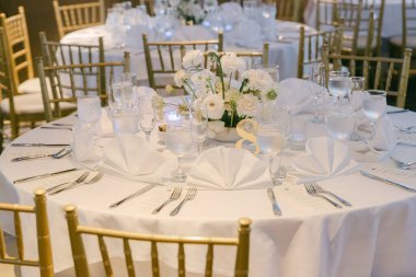 Güzel düzenlenmiş bir yemek masasında beyaz çarşaflar, cam eşyalar ve misafirler için hazır bir çiçek süsü sergilenir..