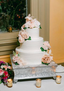 Yumuşak çiçekler ve ılık mumlarla süslenmiş üç katlı güzel bir düğün pastası ortama zarafet katar..