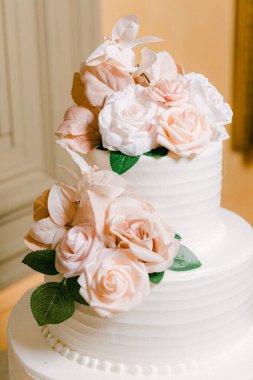 Pembe ve beyaz şeker gülleri ve yeşil yapraklarla süslenmiş üç katlı muhteşem bir düğün pastası. Kutlamalar için ideal..