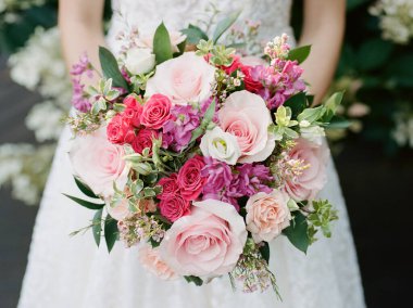 Gelin, bir düğün töreninde yemyeşil bir bahçede canlı pembe ve mor bir çiçek buketi tutuyor..