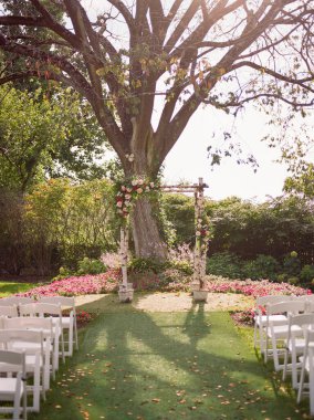 Huzurlu bir düğün töreni çiçek kemerleri ve beyaz sandalyelerle süslü bir bahçede büyük bir ağacın altında hazırlanır..