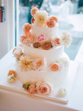 Göz kamaştırıcı, üç katlı bir düğün pastası. Parlak bir resepsiyon alanına yerleştirilmiş çiçeklerle süslenmiş..