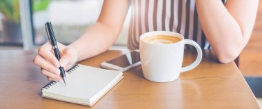 Kadın eli, tahta masadaki bir kalemle bir not defterine yazı yazıyor. Web pankartı..