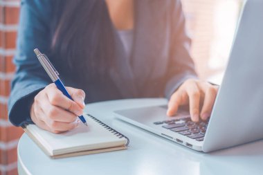 Woman hand is writing on a notepad with a pen and have a laptop computer on the desk in the office.