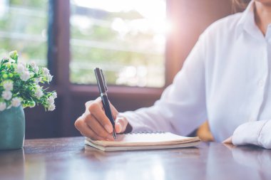 Pencere kenarındaki bir ofiste, tahta bir masanın üzerine siyah bir kalemle not defterine yazan bir kadının elini kapat..