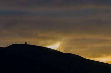 Rivington Pike 'da sonbahar gündoğumu siluet tepe ve güneş bulutları turuncu ve sarı aydınlatıyor.