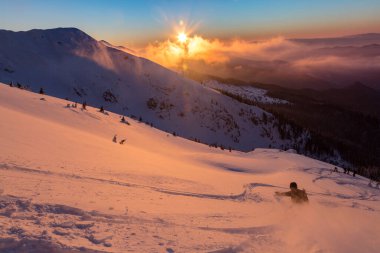 Freeride snowboardcusu, gün batımı turuncu bulutlu gökyüzü ile kaplı güzel, karlı dağlarda, derin karların içinde hareket ediyor. Adrenalin açık hava kış maceraları