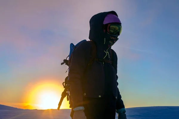 在雪山的背景下 一个戴着面具 头戴安全帽的远足女孩站在太阳圆盘的橙色日出的背景上 一个令人心醉神迷的户外冬季探险 — 图库照片