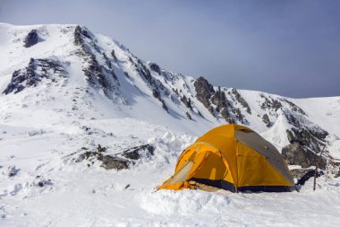 Hikers set orange tent in winter mountains among rocks. Alpinism sport as concept.