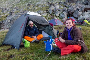 Mountain campsite: Hikers relax by colorful tents, surrounded by rocky terrain and alpine meadows. Couple settles into their tent home clipart