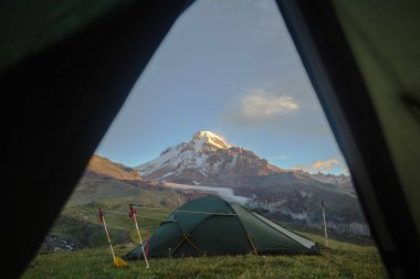 Çadırın girişiyle çerçevelenmiş nefes kesici bir gündoğumu olan Kazbek Dağı 'nın çarpıcı manzarası..