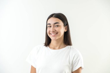 Mutlu ve gülümseyen genç kız stüdyoda kameraya bakıyor. Beyaz arka planda beyaz tişörtle sakin ve kendinden emin.