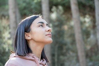 Kısa saçlı ve soluk pembe kazaklı bir kız, ormanın ortasında gökyüzüne bakıyor. Yatay