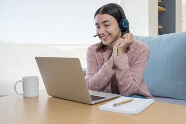 Dizüstü bilgisayarının önünde kulaklıkla video konferansı yapan mutlu kız. O evde bir fincan kahveyle kanepede oturuyor..