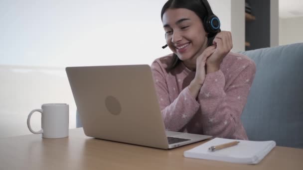 ノートパソコンの前でヘッドフォンで幸せな女の子のビデオ会議 彼女はコーヒーを飲みながらソファの上に座っている — ストック動画