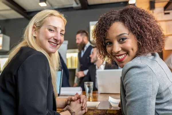Young black south american woman looking at camera with her blonde caucasian colleague while working in modern office - Break from work and coworking concept