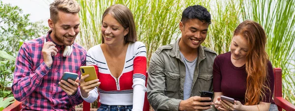 大学のキャンパス内のデザートガーデンで一緒にスマートフォンを見て楽しんでいる多文化の友人との水平バナーやヘッダー 携帯電話に依存している人々 — ストック写真