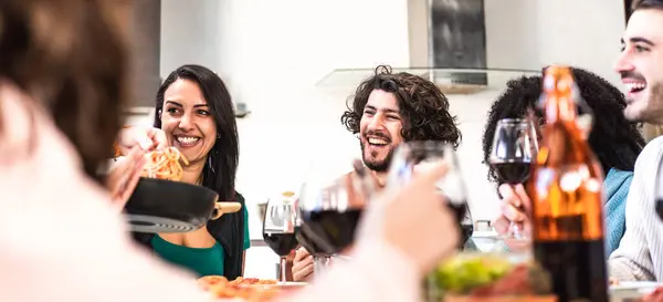 Friends toasting red wine glasses at dinner while eating spaghetti with tomato - Multiethnic guys having fun together at dinner with italian pasta and red wine - Banner or header with bright filter