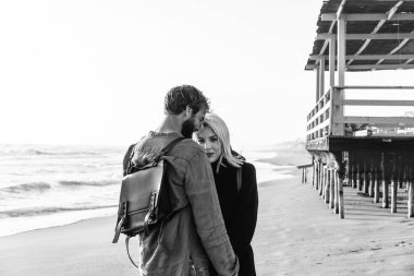 Romantik hippi çift, sahilde, ahşap yapının yanında duruyor. Erkek arkadaş ve kız arkadaş, güzel kıyafetler ve klasik sırt çantaları ile denizin önünde duruyorlar.