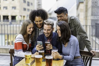 Bir grup farklı genç arkadaş açık hava barında bira içerken bir akıllı telefona gülüp bakıyorlar. Kentsel bir sosyal ortamda eğlenceli bir bağ ve neşe paylaşıyorlar..