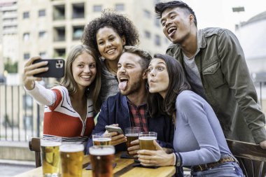 Bir grup farklı genç arkadaş bir barda selfie çekerken ve bira içerken komik surat ifadeleri takınıyorlar. Rahat bir şehir ortamında gülüyor ve arkadaşlığı kutluyorlar. Gen Z yaşam tarzı konsepti.