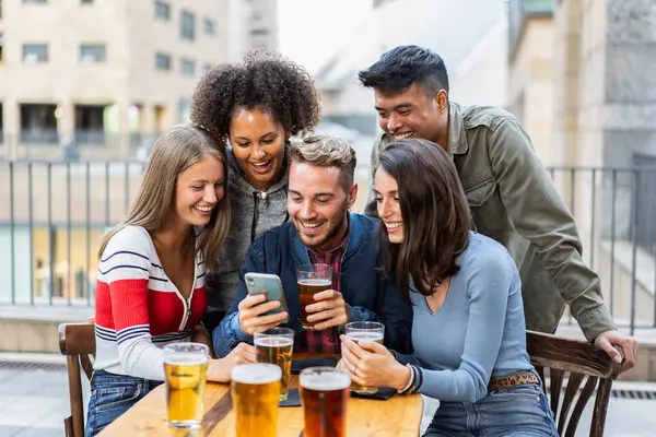 Bir grup farklı genç arkadaş açık hava barında bira içerken bir akıllı telefona gülüp bakıyorlar. Kentsel bir sosyal ortamda eğlenceli bir bağ ve neşe paylaşıyorlar..
