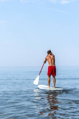 Genç adam yazın açık mavi gökyüzünün altında sakin denizde kürek çekiyor. Asgari arkaplan ve canlı renk karşıtlığına sahip aktif yaşam tarzı ve su sporu kavramı.