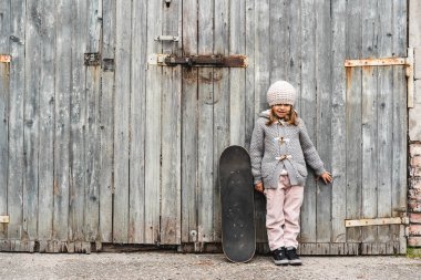 Kışlık yün elbiseli, kaykaylı şirin küçük bir kız grunge ahşap arka planda dururken - aktivite kavramı ve çocukluk - avluda açık hava oyunları - tanıtım alanı