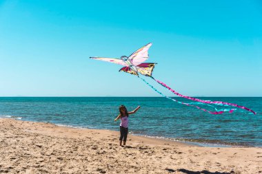 Denizin üzerinde renkli bir kelebek uçurtması uçururken kumsalda oynayan gülümseyen küçük kız. Suyun kenarında neşeli ve tasasız bir çocukluk anı. Açık hava aktivitesi ve yaz özgürlüğü kavramı.