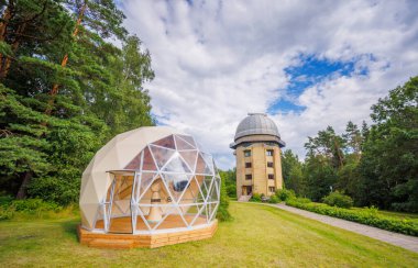 Ağaçlarla çevrili kubbeleri ve bulutlu gökyüzünün altındaki yeşil çimlerle gözlemevi manzarası.
