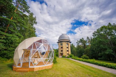 Ağaçlarla çevrili kubbeleri ve bulutlu gökyüzünün altındaki yeşil çimlerle gözlemevi manzarası.