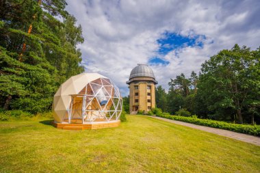 Ağaçlarla çevrili kubbeleri ve bulutlu gökyüzünün altındaki yeşil çimlerle gözlemevi manzarası.