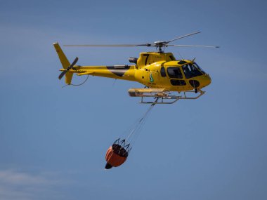 Plasencia 'da. İSPAN. - 12 Eylül 2024: Sarı itfaiye helikopteri açık mavi gökyüzüne doğru uçarken elinde bir kova suyla.