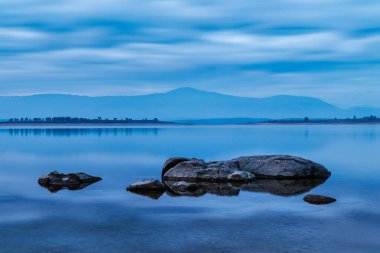 Durgun göl ve kayalar mavi bulutlu alacakaranlık gökyüzünün altında.
