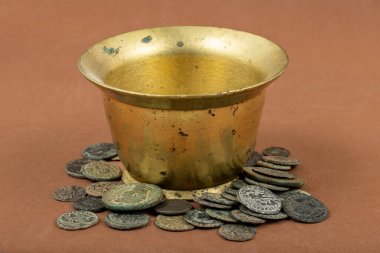 Kahverengi arka planda antik Roma sikkeleri ile çevrili eski pirinç bardak.