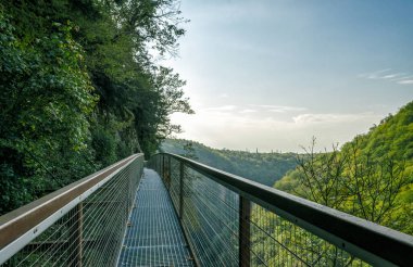 Gürcistan 'da bir kanyonda, dağların, ormanın ve bulutlu gökyüzünün arka planına karşı uzun ve güvenli bir turistik metal köprü.