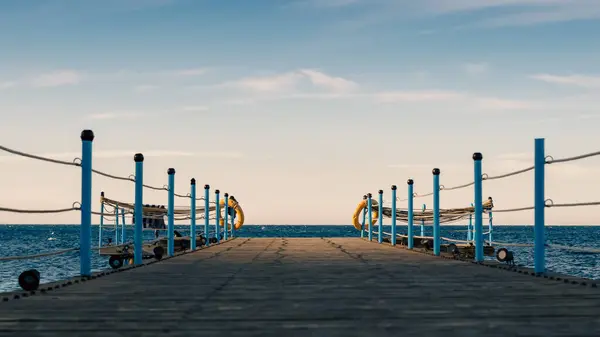 stock image wooden platform with blue posts with ropes and orange lifebuoys on the background of the sea and sky with clouds