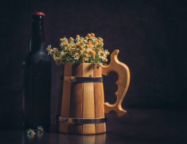 Bir şişe birayla, tahta bardakla ve koyu kahverengi arka planda köpük taklidi yapan papatyalarla dolu bir hayat.