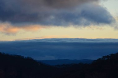 Kara fırtına bulutları ufku dolduruyor. Gökyüzündeki diğer bulutlara günbatımının renkleri değmiştir. Ormanla kaplı tepeler ön planda..