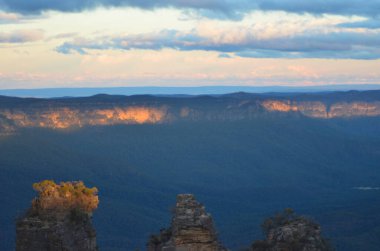 Günbatımının ışığı bulutların arasından geçip Avustralya 'nın Mavi Dağları' ndaki en uzun üç kız kardeşin tepesine ulaşıyor. Uzak dağ sırasının tepesini de renklendirir. İkisinin arasında ormanla dolu gölgeli bir vadi var..