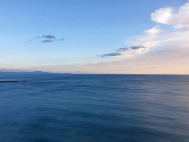 Sakin bir okyanus ufka kadar uzanıyor. Dalgakıran bir tarafta, uzak tepelerin çerçevelediği bir tekne var. Su koyu mavi ve bulutlar gün batımının renklerine sahip..