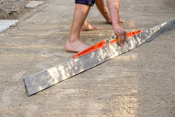 A spirit level in man\'s hands. A barefoot man levels the sandy platform with an aluminum bar. Evening golden hour. Domestic life. Copy space.