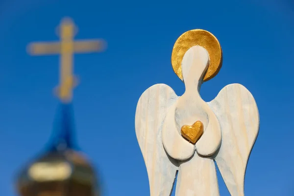 Wooden angel with a golden halo and heart in his hands on a blurred church background. Blue sky.