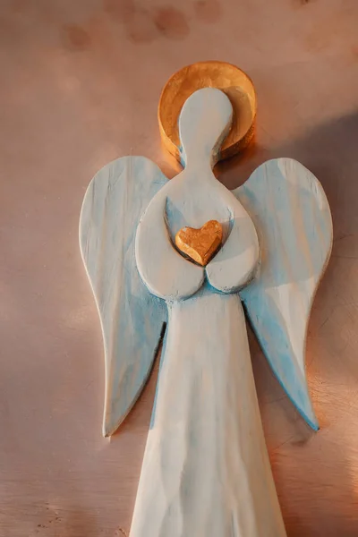 A wooden figure of an Angel with a golden halo. Golden heart. Peach fuzz color background.