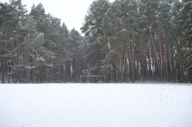 Uzun çam ağaçlarından oluşan sık bir ormanla çevrili karla kaplı futbol sahası.