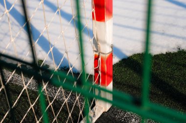 Futbol kaleleri kışın suni bir çim sahasına kırmızı ve beyaza boyanmış. Suni çayırda kar. Güneş ışığı. Keskin gölge.