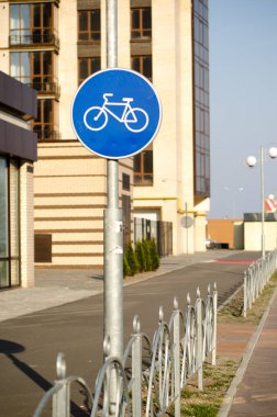 Şehirde yüksek bir binanın yanında metal çitli mavi yol tabelası var.