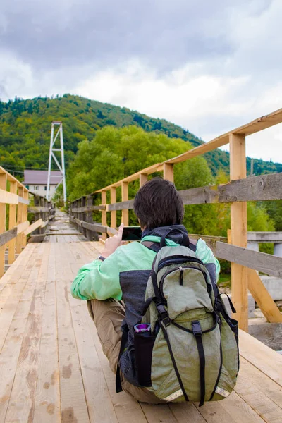 緑のジャケットとリュックを着た若者が山の近くの木製の橋の上に座り スマートフォンで写真を撮る — ストック写真